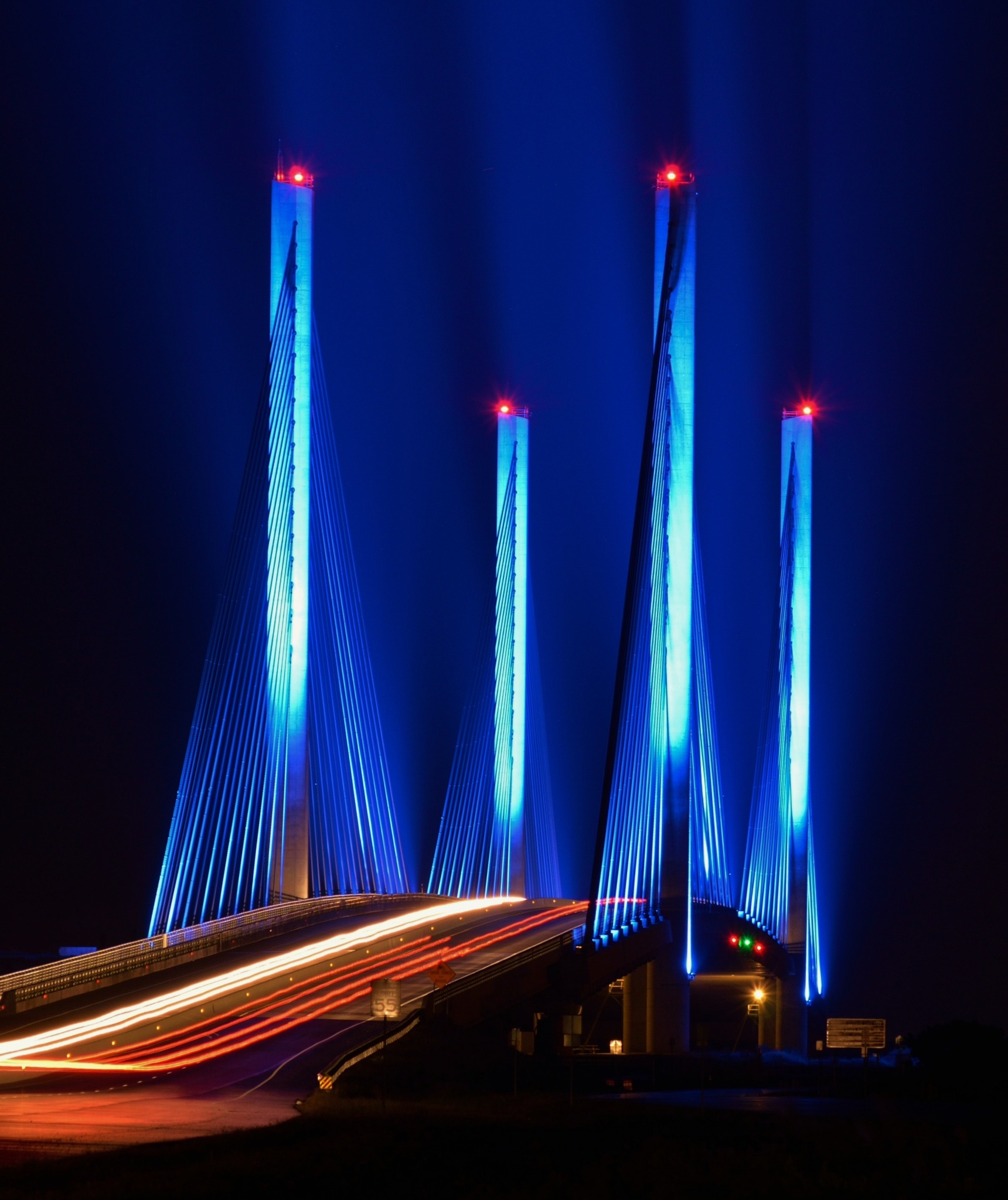 The Charles W. Cullen Bridge illuminated blue at night, with four towers and white and red long-exposure traffic trails crossing the Indian River Inlet.