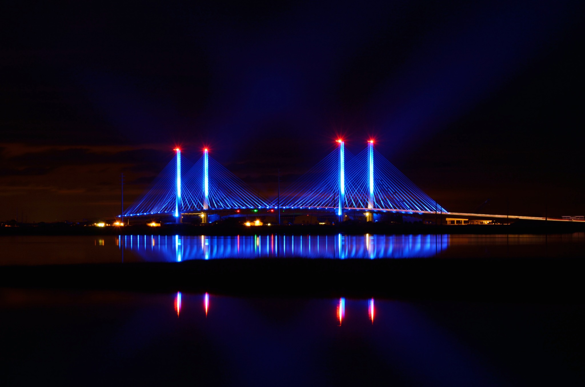 The Charles W. Cullen Bridge illuminated blue at night in a wide view, with its four towers and colored lights reflected in the calm water of Indian River Inlet.